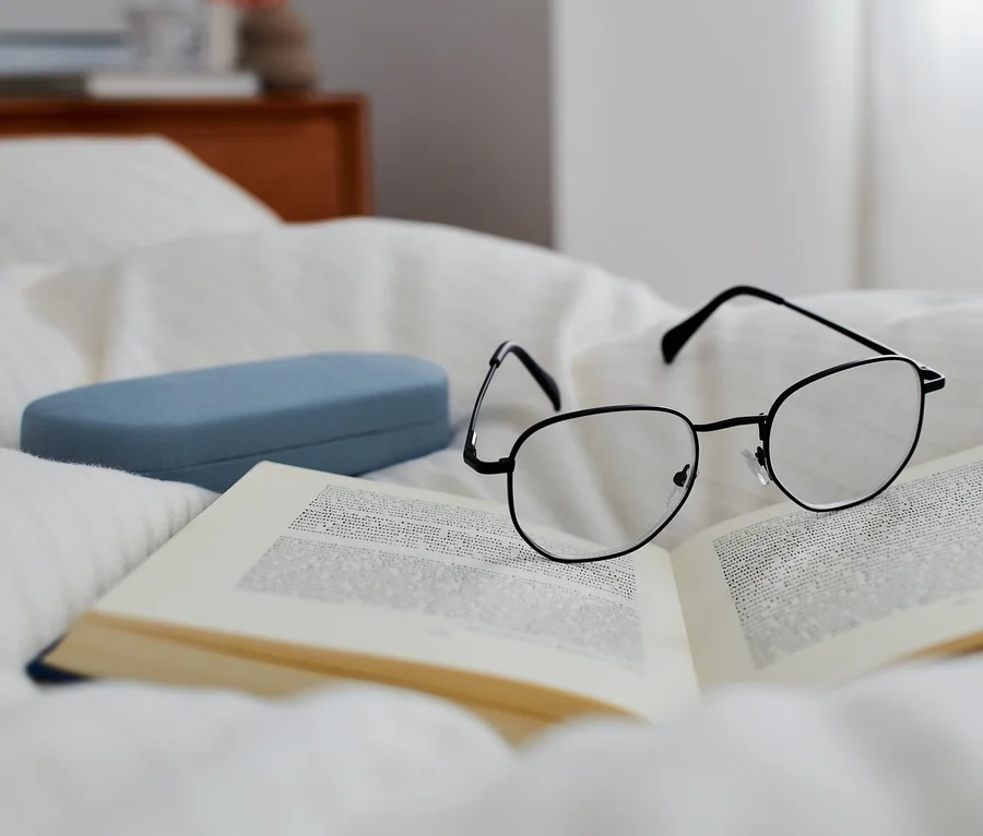 Eine Lesebrille mit Etui liegt auf einem offenen Buch auf weißer Bettwäsche.