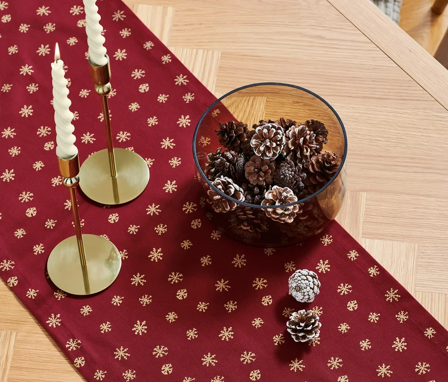 Auf einem Holztisch liegt ein roter Tischläufer mit goldenen Schneeflocken, auf dem zwei goldene Kerzenständer mit weißen Kerzen und eine Glasschale voller Tannenzapfen stehen.