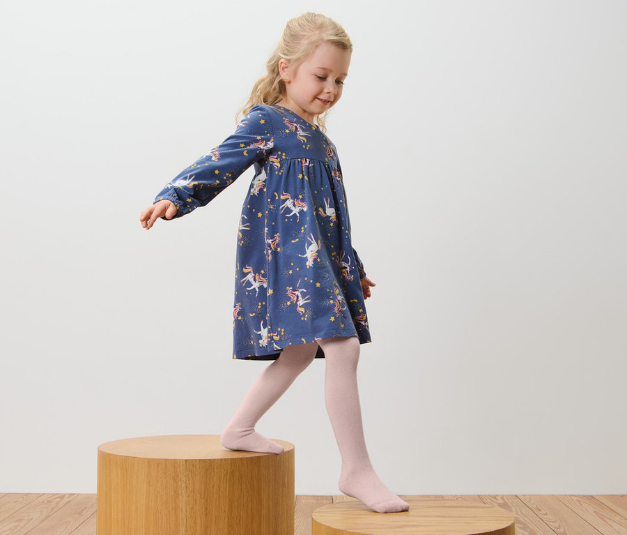 Ein Mädchen in einem blauen Kleid mit Einhorn-Print und rosa Baumwoll-Strumpfhosen, 5er-Pack, rosa-braun, geht auf Holzstufen.