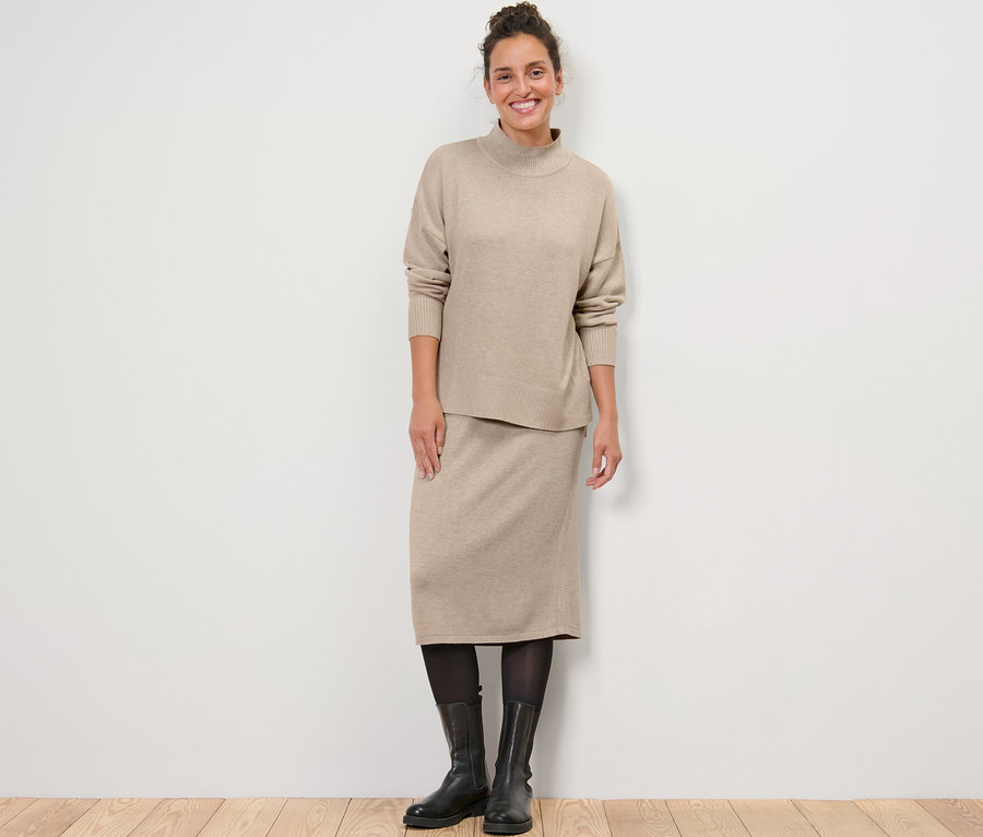 Eine Frau steht und trägt einen Feinstrickpullover mit Stehkragen und einen Strickrock.