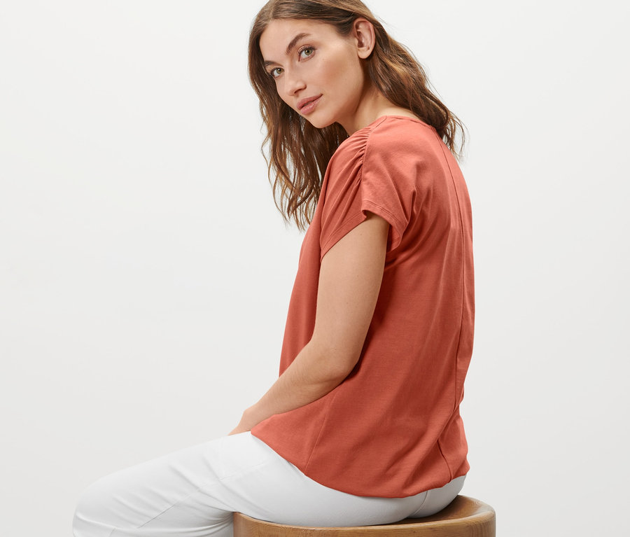 Frau sitzt auf einem Hocker und trägt ein apricotfarbenes Blusenshirt.