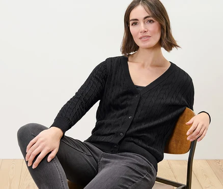 Frau sitzt auf einem Stuhl und trägt einen schwarzen Feinstrickcardigan mit Zopfmuster.