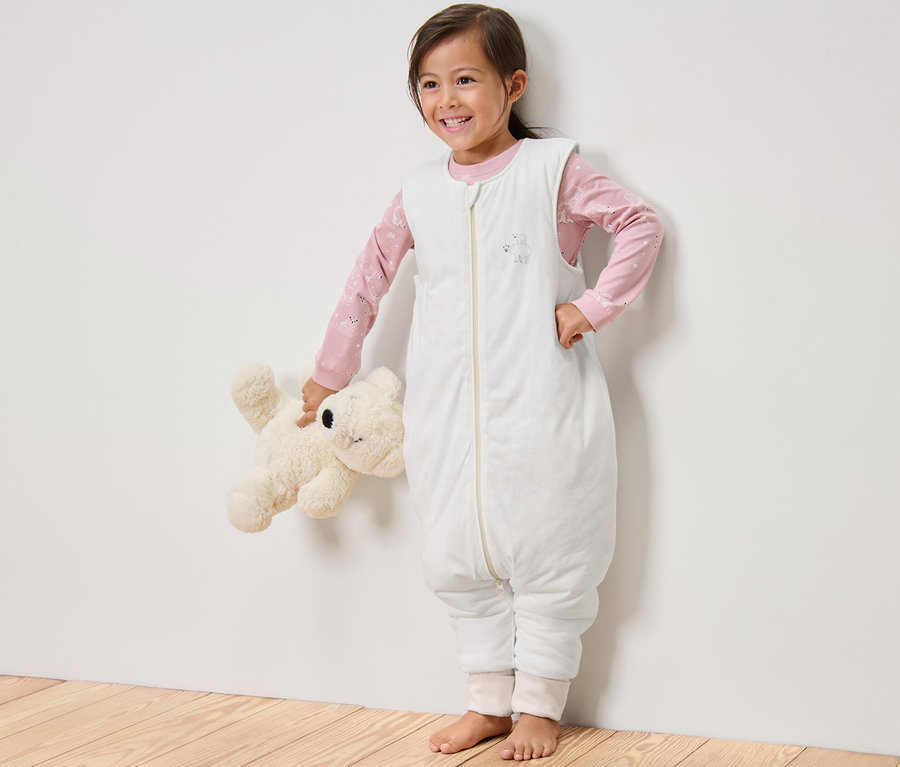 Ein lächelndes Mädchen steht und hält einen Teddybären. Es trägt einen Schlafsack mit Beinen, 2,5 TOG, 110 cm.