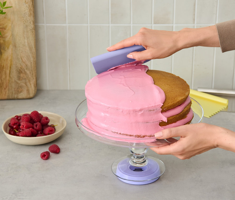 Person trägt mit einem lila Werkzeug rosa Zuckerguss auf eine Torte auf, die auf einer Torten-Drehscheibe steht.