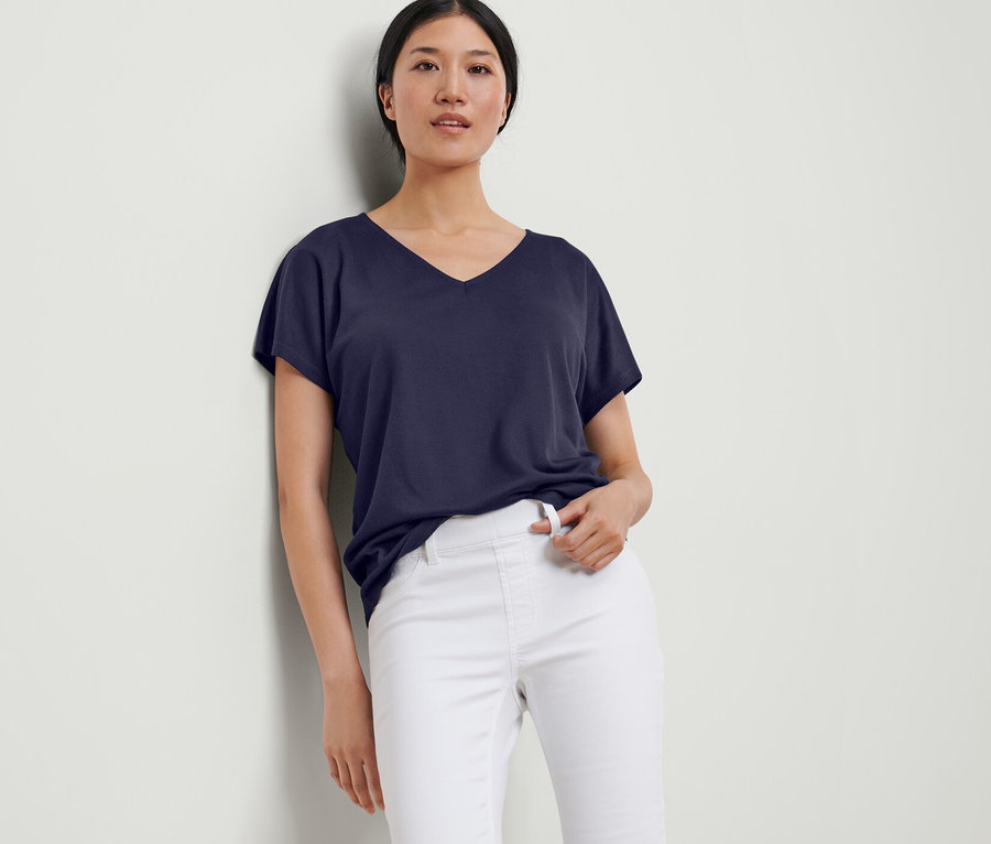 Eine Frau lehnt an einer Wand und trägt ein dunkelblaues, strukturiertes Shirt und eine weiße Hose.