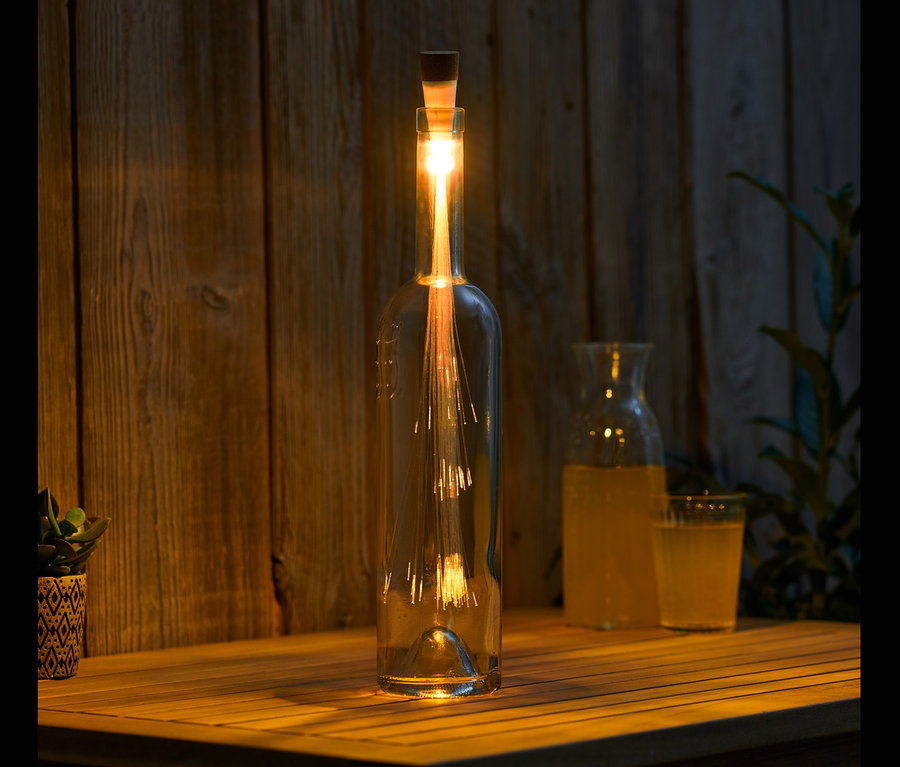 Auf einem Holztisch steht eine Flasche mit einem LED-Korken mit Akku, daneben ein Krug und ein Glas mit Getränk.