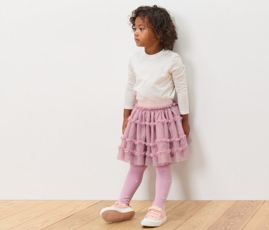 Mädchen trägt einen Kinder-Tüllrock, ein Langarmshirt, Leggings und Turnschuhe.