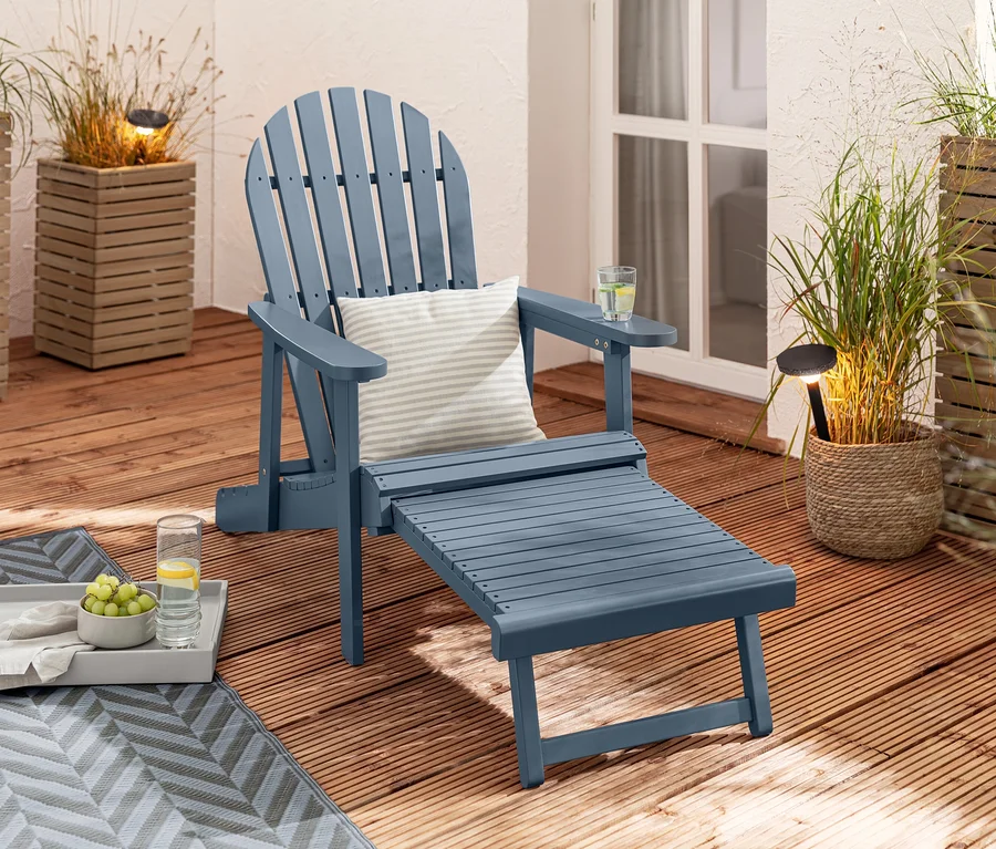 Ein blauer Single Adirondack Relax-Sessel mit ausziehbarer Fußlehne steht auf einer Holzterrasse neben 2 Solar-Steckleuchten und 2 Outdoor-Kissen in Leinen-Haptik.