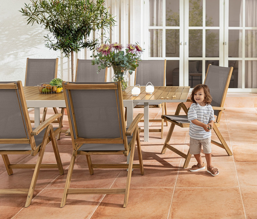 Ein Junge steht neben einem Ausziehtisch »Liska« aus Aluminium und einem Hochlehner »Lenja« mit Holzrahmen. Im Hintergrund befinden sich ein Barwagen und ein Akku-Tischleuchten-Set.