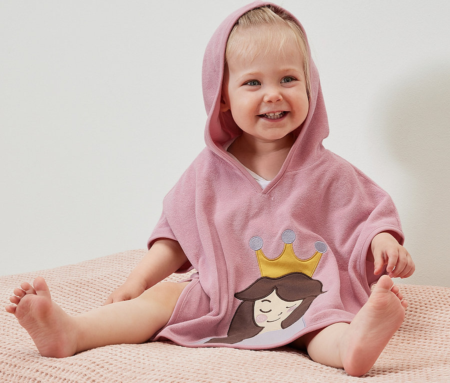Ein lächelndes Kind sitzt auf einer rosafarbenen Oberfläche und trägt einen Frottierponcho mit Kapuze und einem Prinzessinenmotiv.
