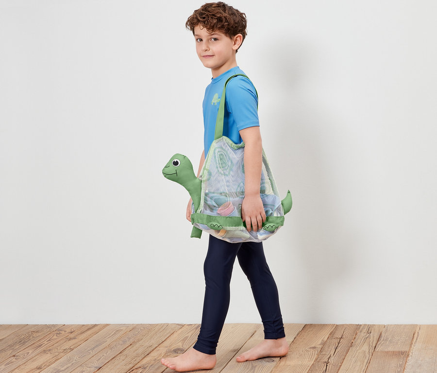 Ein Junge geht und trägt eine Strandspielzeug-Netztasche mit Schildkrötenmotiv.