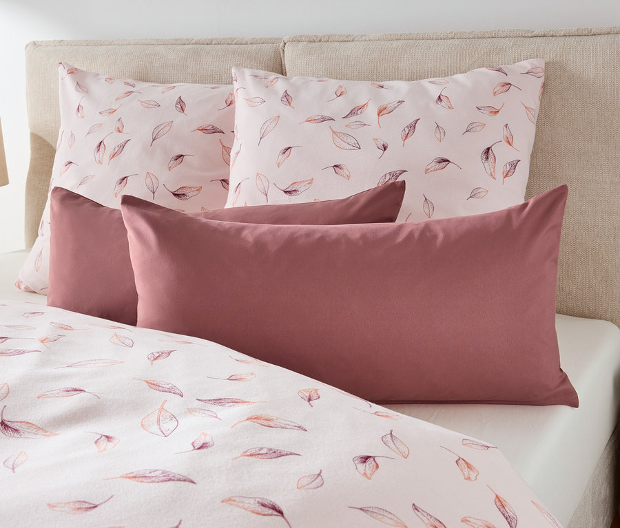 Auf einem Bett liegen zwei Kissen mit Blätterdruck und zwei kastanienbraune Kissen.