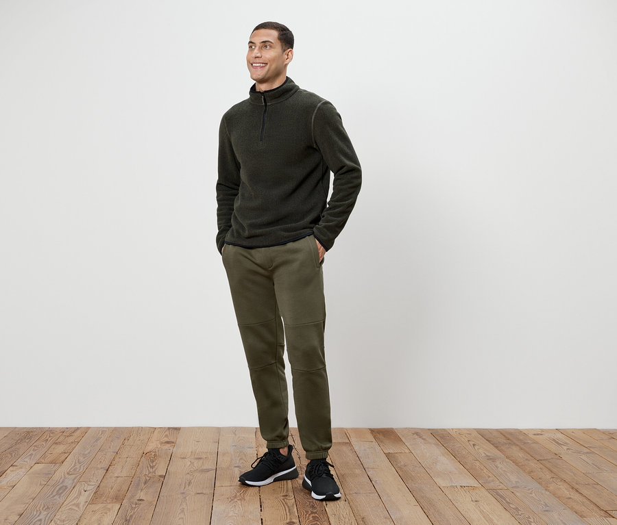 Ein Mann in olivgrünen Hosen und einem dunkelgrauen Fleece-Pullover mit Reißverschluss steht auf einem Holzboden.