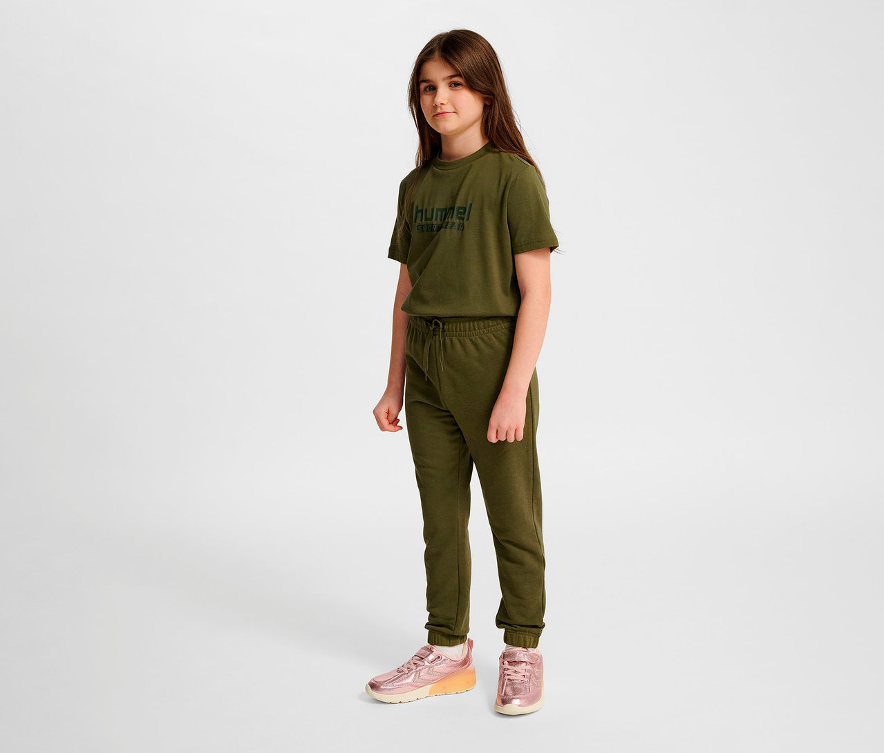 Mädchen steht in grünem T-Shirt, grüner HUMMEL HMLJR Base Regular Pants und pinken Schuhen.