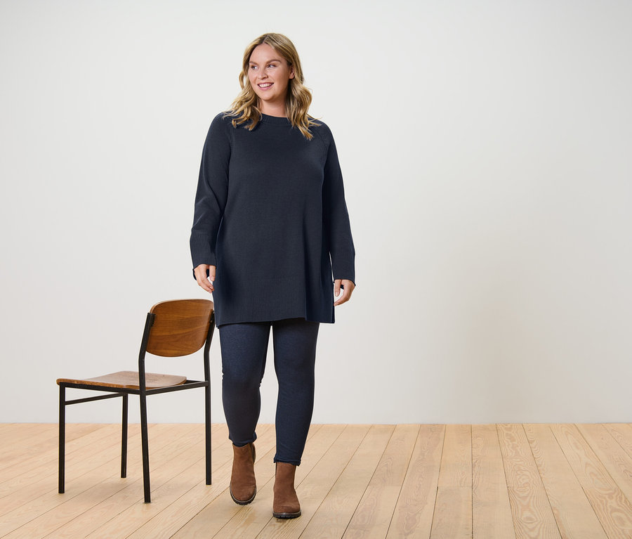 Eine Frau trägt einen langen, dunkelbraunen Strickpullover und gefütterte Leggings in Jeansoptik und geht.