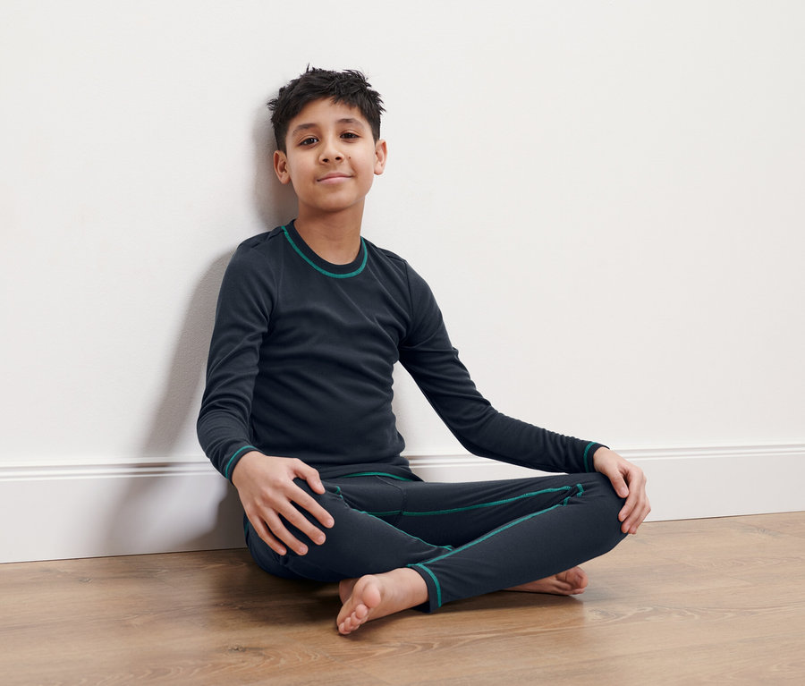 Ein Junge sitzt mit gekreuzten Beinen auf dem Boden und lehnt sich an eine weiße Wand. Er trägt dunkelblaue Thermounterwäsche.