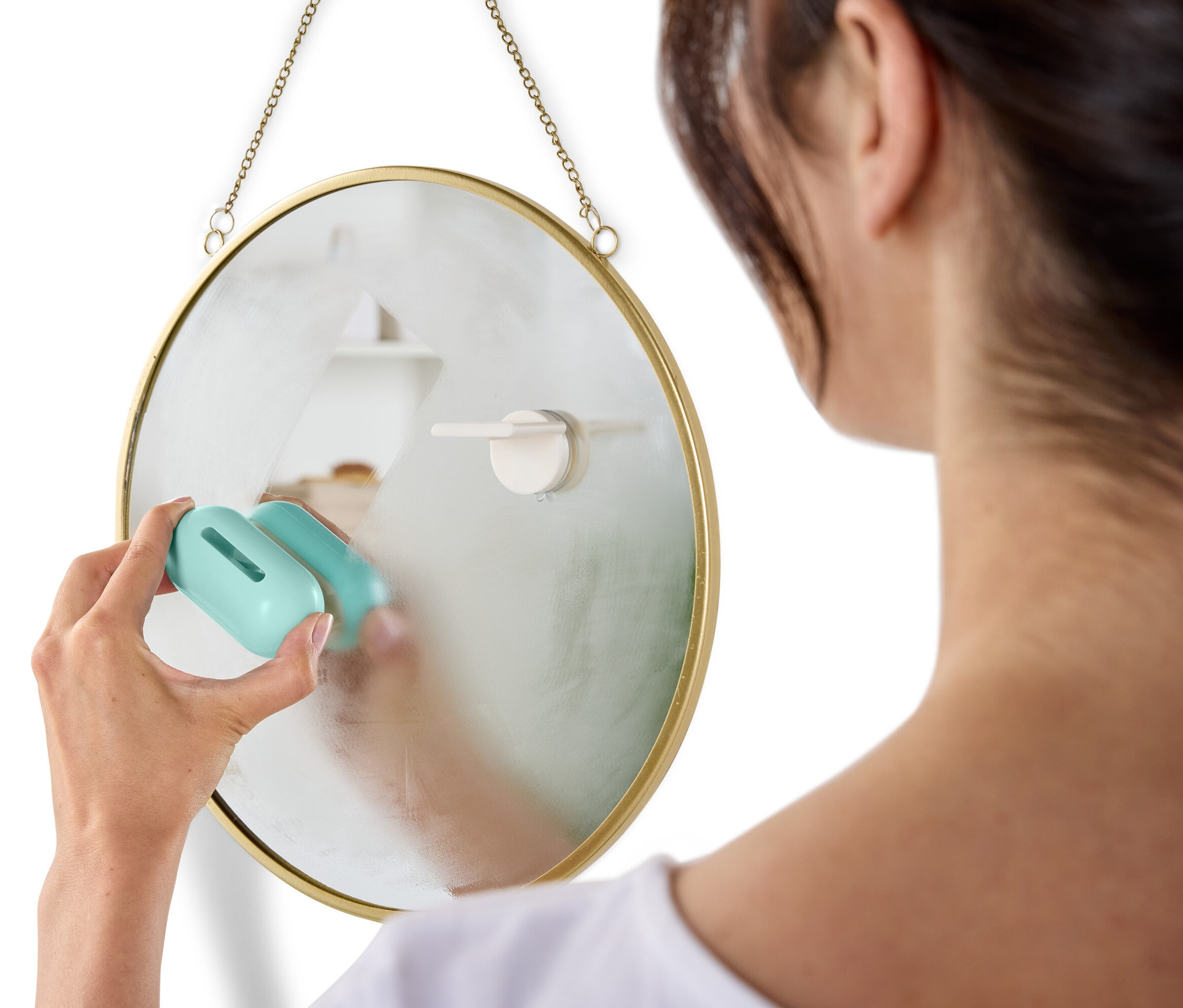 Frau putzt einen runden Spiegel, der mit einer Kette an der Wand hängt, mit einem blauen Putzmittel.