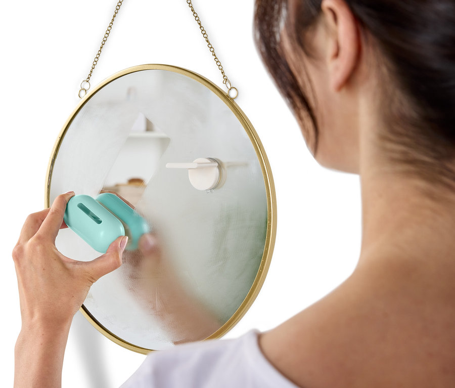 Frau putzt einen runden Spiegel, der mit einer Kette an der Wand hängt, mit einem blauen Putzmittel.