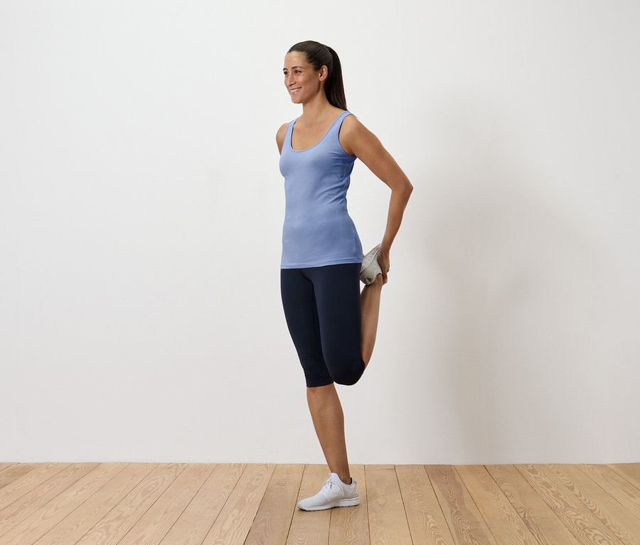 Frau in blauem Tanktop und dunklen Leggings dehnt ihren Quadrizeps, indem sie ihren Fuß hinter sich hält.
