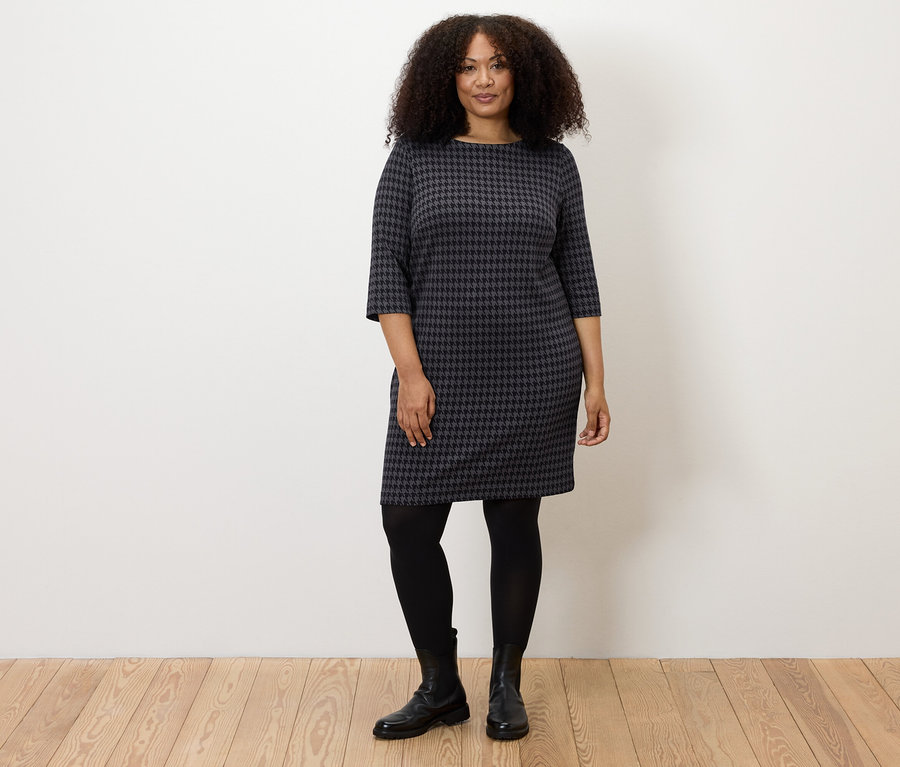 Frau steht und trägt ein Jerseykleid mit Hahnentritt-Jacquard, eine schwarze Strumpfhose und schwarze Stiefel.