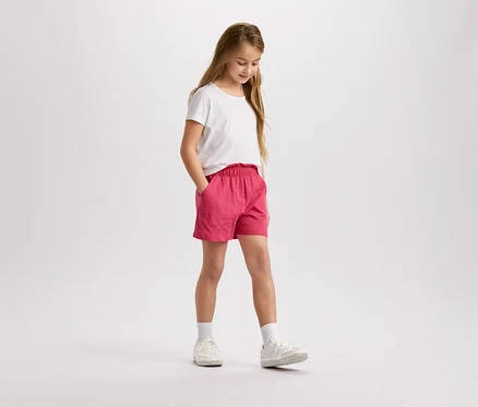 Mädchen mit blonden Haaren, weißem T-Shirt und pinkfarbenen Shorts steht da und schaut nach unten.
