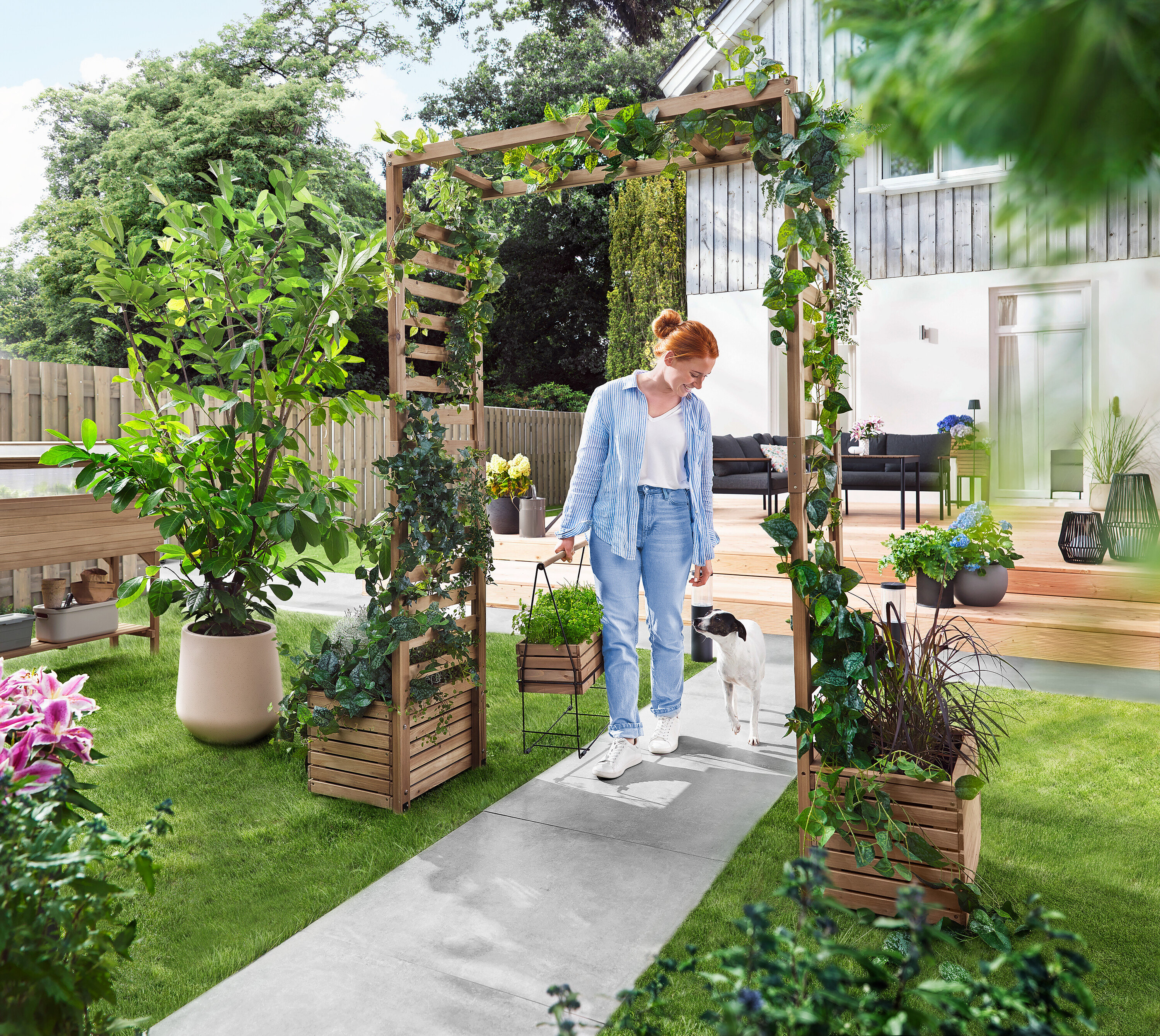 Eine Frau geht mit einem Hund durch einen Gartenbogen, der mit Pflanzen bewachsen ist. Sie trägt eine Pflanzbox.