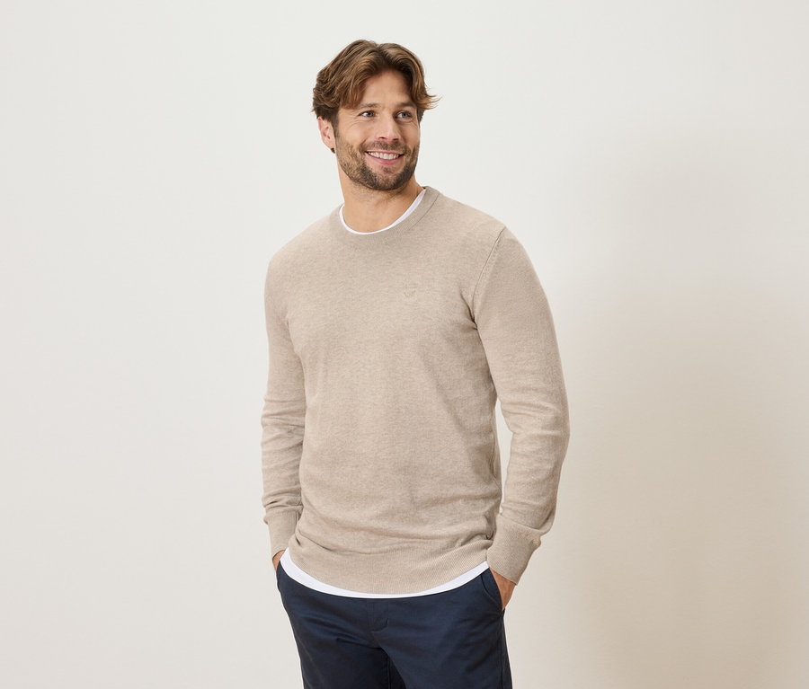 Mann mit beigem Pullover und blauen Hosen, die Hände in den Hosentaschen, lächelnd und seitlich blickend.