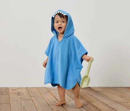 Ein Kleinkind trägt einen blauen Hai-Poncho und hält eine grüne Schaufel in der Hand.