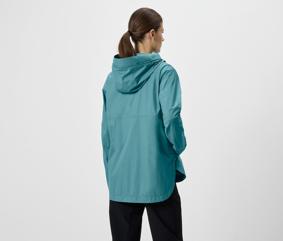 Rückansicht einer Frau mit Pferdeschwanz in einer türkisfarbenen Regenjacke mit Kapuze und schwarzen Hosen vor einem weißen Hintergrund.