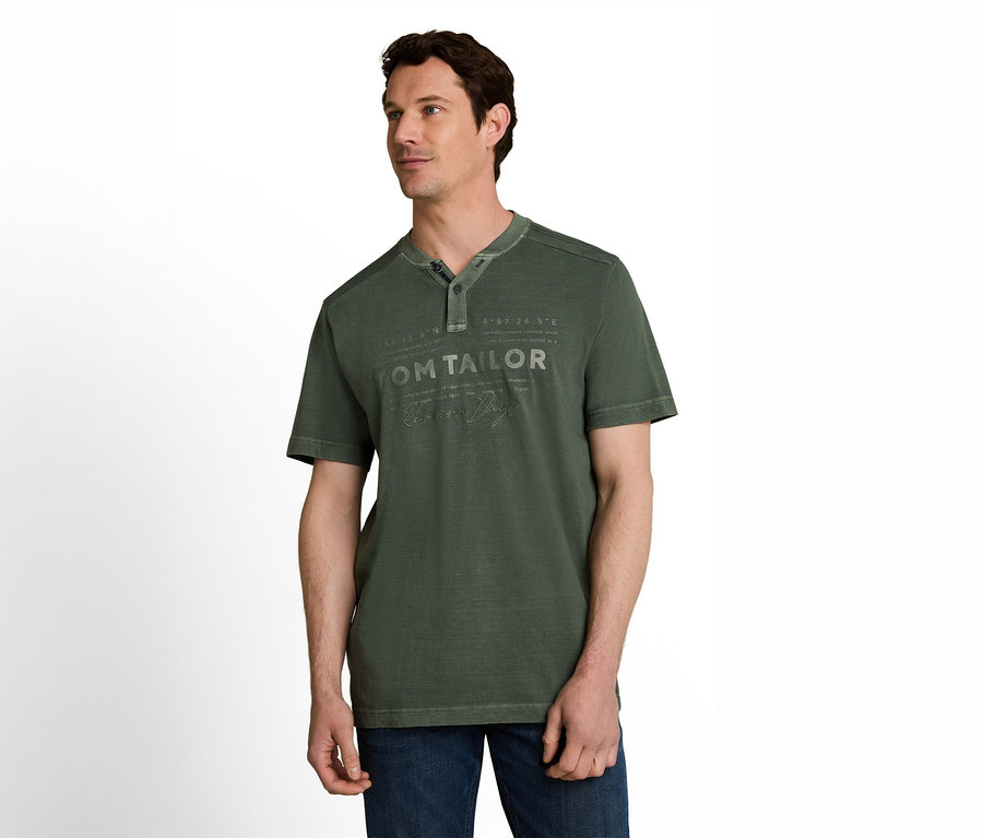 Ein Mann mit dunklem Haar trägt ein grünes Henley-T-Shirt mit "TOM TAILOR"-Aufschrift und blaue Jeans vor weißem Hintergrund.