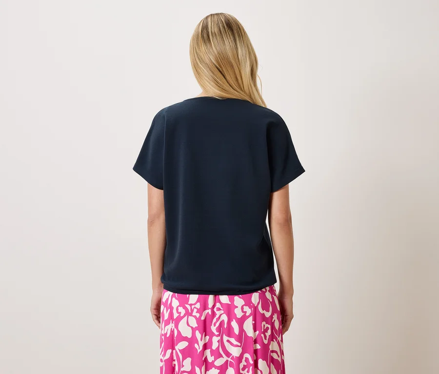 Rückenansicht einer Frau mit blonden Haaren, die ein marineblaues T-Shirt und einen pinkfarbenen Rock mit Blumendruck trägt.