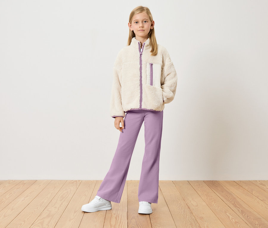 Mädchen mit beiger Teddyjacke mit lila Details, lila Hose und weißen Turnschuhen steht vor einer weißen Wand.
