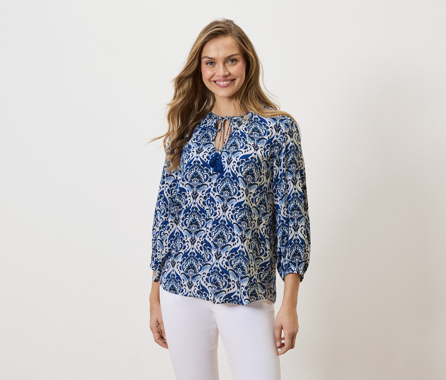 Frau mit langen blonden Haaren, die eine blau-weiß gemusterte Bluse und eine weiße Hose trägt und lächelt.