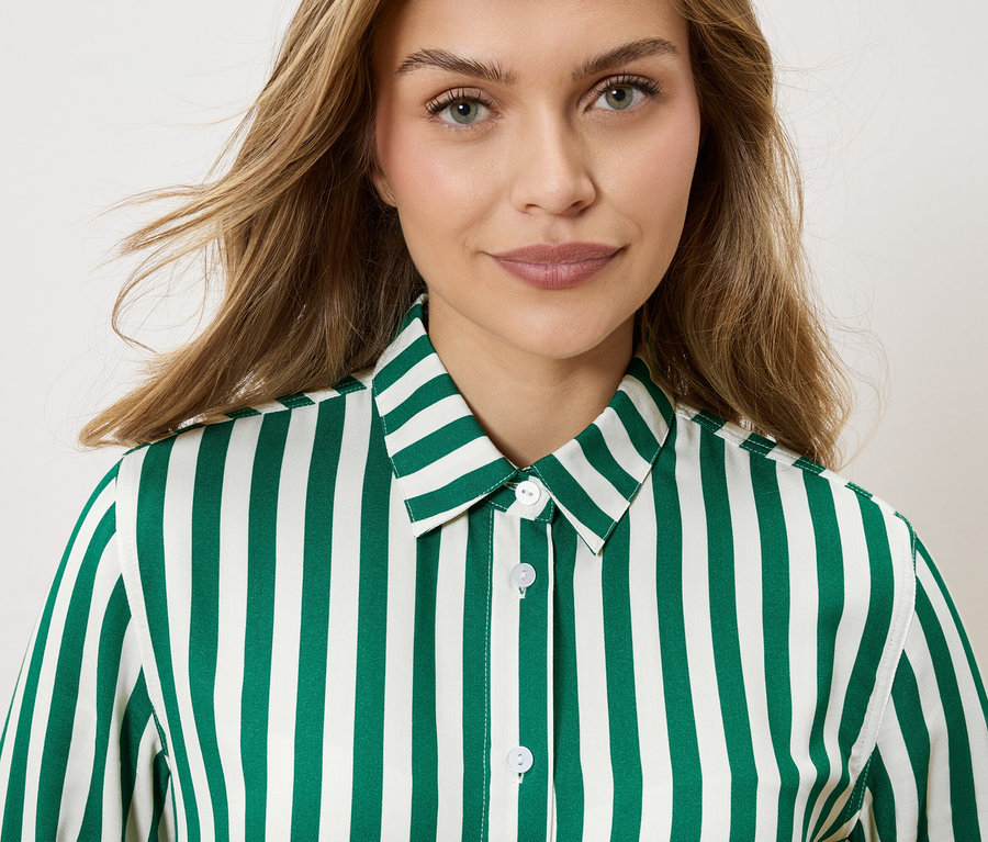 Porträt einer Frau mit blonden, gewellten Haaren, die ein gestreiftes, smaragdgrünes und weißes Hemd trägt.