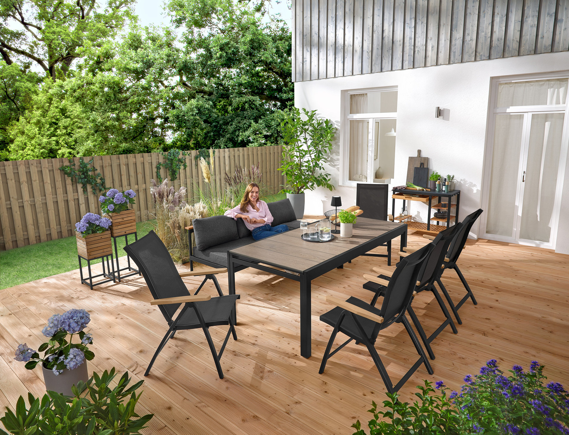Eine Frau sitzt auf einer 2-in-1-Gartenbank ‒ Sonnenliege »Elin«. Im Hintergrund stehen 2 Pflanzsäulen »Elin«. Auf der Terrasse stehen auch ein Grillwagen mit Polyzement-Arbeitsfläche und ein ausziehbarer Gartentisch mit Duraboard-Tischplatte.