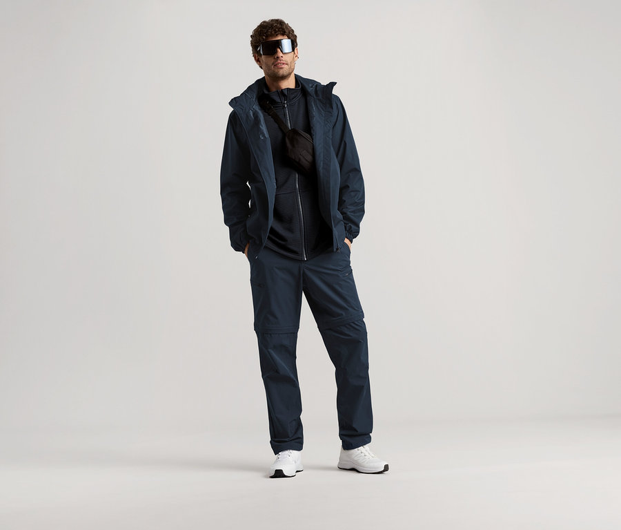 Mann steht mit Sonnenbrille, marineblauer Jacke, Hose und Bauchtasche vor hellem Hintergrund.