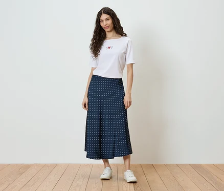 Frau mit lockigem Haar trägt ein weißes T-Shirt mit Herz-Motiv und einen dunkelblauen Rock mit weißen Punkten, dazu weiße Turnschuhe.
