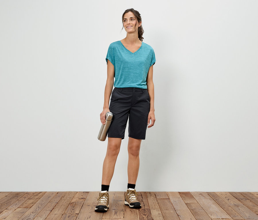 Frau steht vor einer weißen Wand, hält eine silberne Flasche, trägt ein blaues T-Shirt, Shorts und Wanderschuhe.