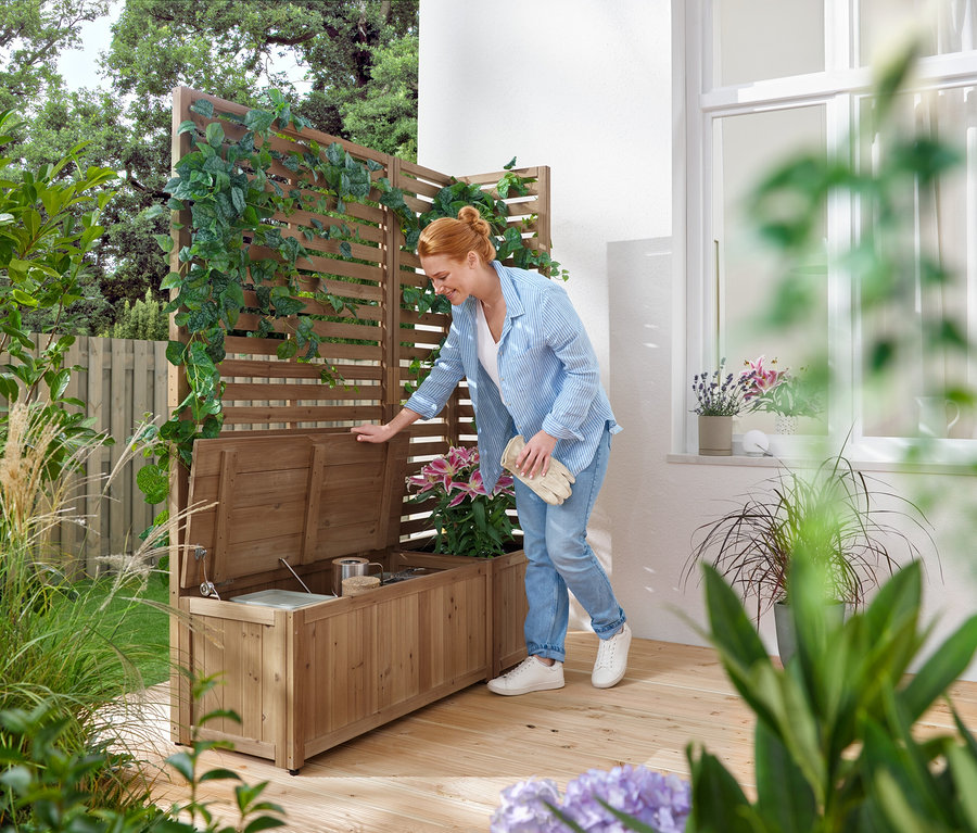 Frau öffnet eine hölzerne Gartenbank mit Stauraum, neben einem Holzzaun mit Kletterpflanzen.