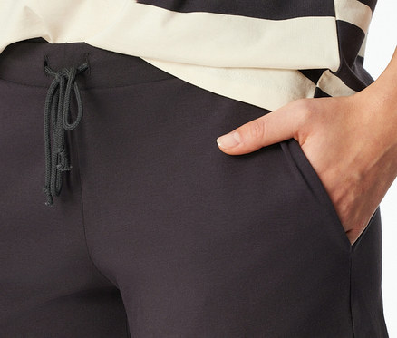 Nahaufnahme einer Person in einer dunkelgrauen SCHIESSER Shorty-Set Pyjamahose mit Kordelzug und einer Hand in der rechten Tasche. Ein schwarz-cremefarbenes Ringelshirt ist über der Hose sichtbar.