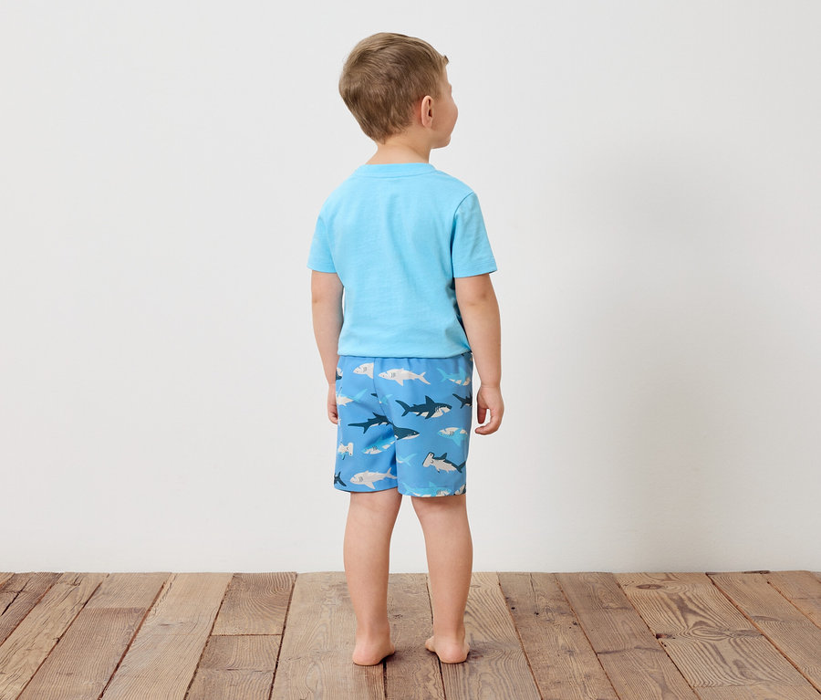 Rückansicht eines Jungen mit hellblauem T-Shirt und Badehose mit Hai-Motiv, stehend auf Holzboden vor einer weißen Wand.