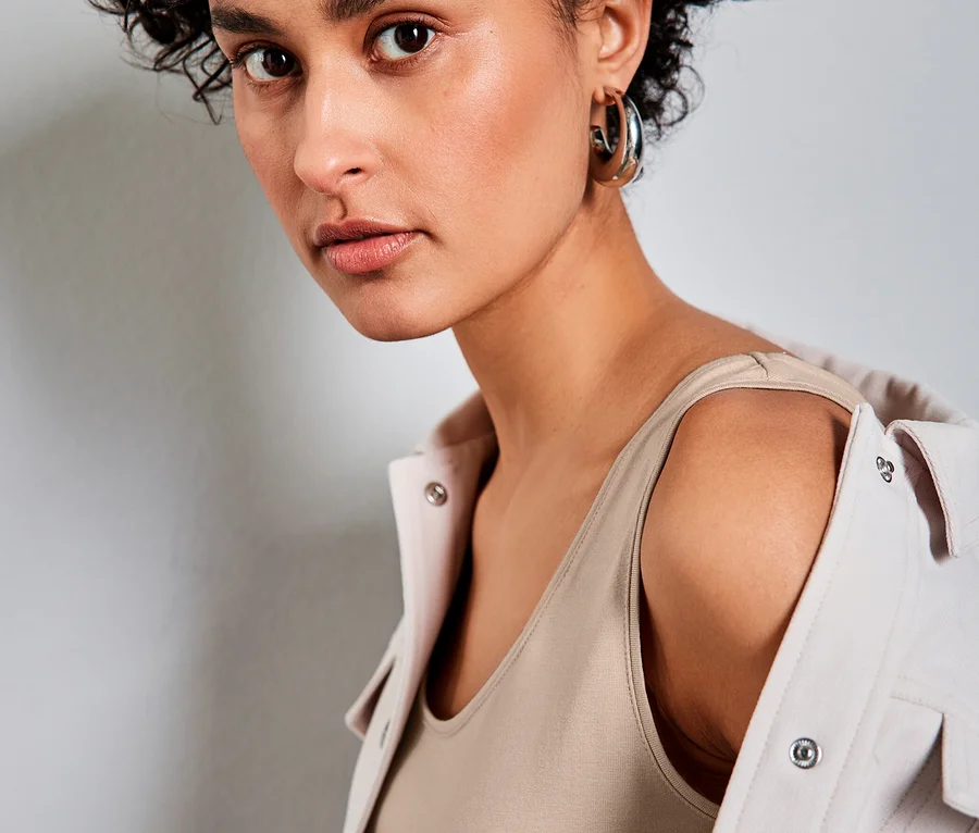 Frau mit kurzen Locken trägt ein beiges Street One Basic-Top und eine helle Jacke über den Schultern, blickt in die Kamera.