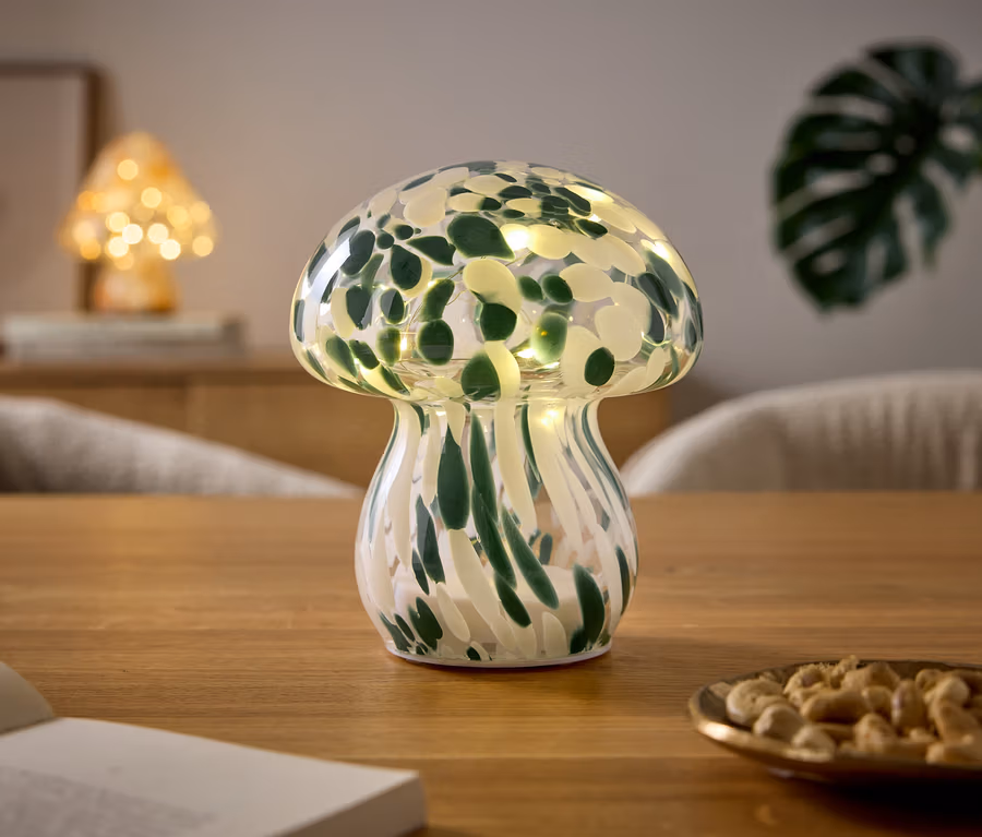 Pilzförmige Lampe mit grün-weißem Muster und Lichtern steht auf einem Holztisch. Daneben liegen ein offenes Buch und eine Schale mit Cashewkernen. Im Hintergrund ist eine weitere Pilzlampe und eine Pflanze zu sehen.