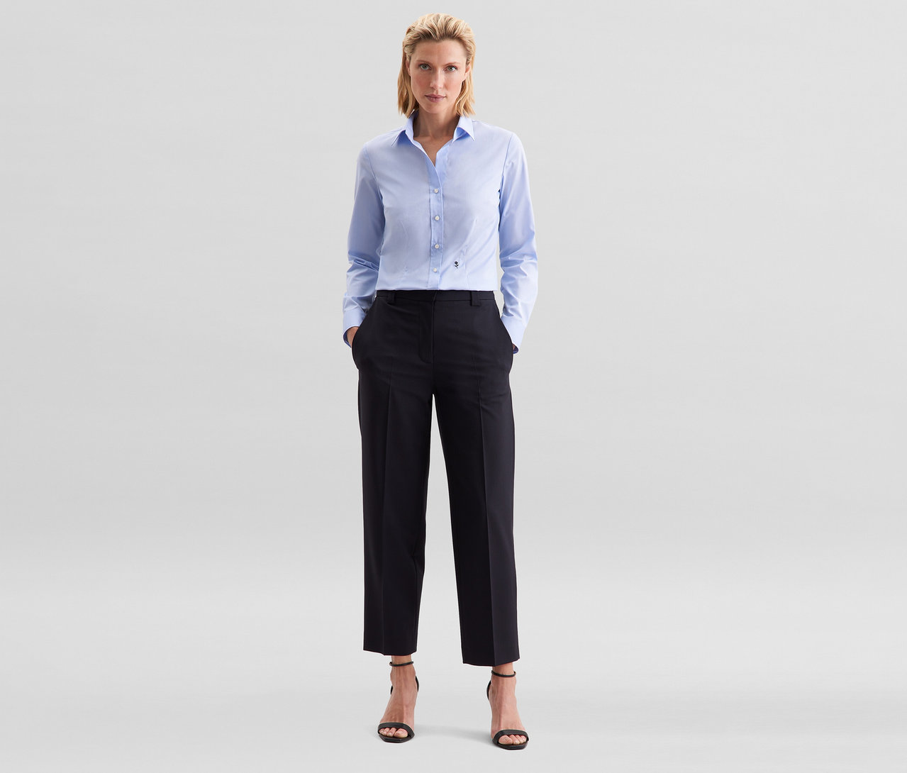 Frau steht mit Händen in den Taschen, trägt eine hellblaue Seidensticker Bluse Slim Fit non iron, schwarze Hose und schwarze Sandaletten.