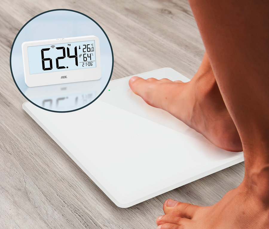 Füße stehen auf einer weißen digitalen Personenwaage, die 62,4 kg, 26,0 °C, 64 % Luftfeuchtigkeit und das Datum 21-06 anzeigt.