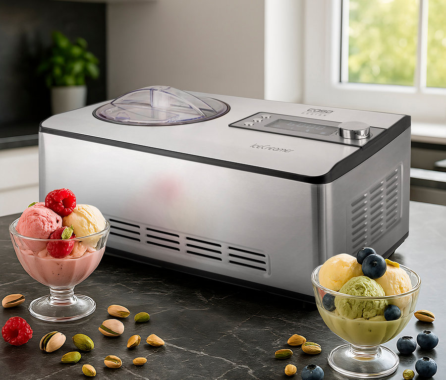 Silberne Eismaschine auf dunkler Arbeitsplatte, daneben zwei Glasschalen mit Eiscreme. Eine Schale enthält rosa und gelbes Eis mit Himbeeren und Pistazien, die andere grünes und gelbes Eis mit Blaubeeren und Pistazien. Beeren und Pistazien liegen verstreut.