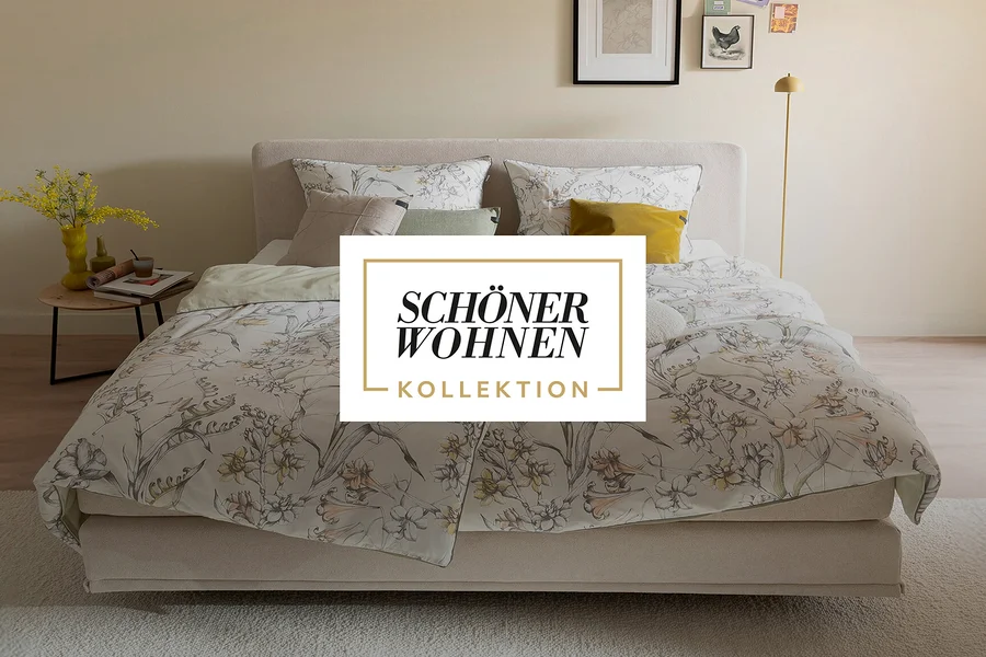 Ein Bett mit hellbeigem Kopfteil und floraler Bettwäsche in Hellgrün-, Gelb- und Brauntönen. Mehrere Kissen, einige passend zur Bettwäsche, andere in Beige, Hellgrün und Gelb, liegen auf dem Bett. Ein Holznachttisch links trägt eine gelbe Vase mit Blumen, eine grüne Vase, Bücher und eine Tasse. Eine gelbe Stehlampe steht rechts neben dem Bett.