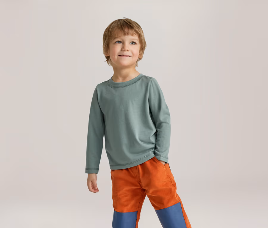 Ein kleiner Junge mit hellbraunen Haaren trägt ein grünes Langarmshirt und orangefarbene Hosen mit blauen Knieflicken. Er steht lächelnd da und blickt nach oben.