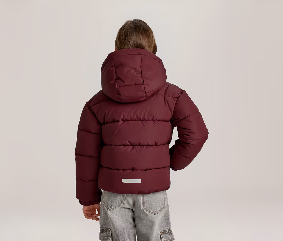Kind von hinten in einer bordeauxroten Steppjacke mit Kapuze und hellgrauen Cargohosen. Auf dem Rücken der Jacke befindet sich ein reflektierender Streifen.