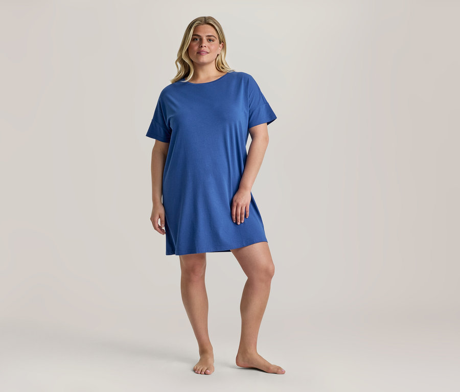 Blonde Frau steht barfuß und trägt ein blaues Nachthemd mit kurzen Ärmeln, das bis über die Knie reicht.